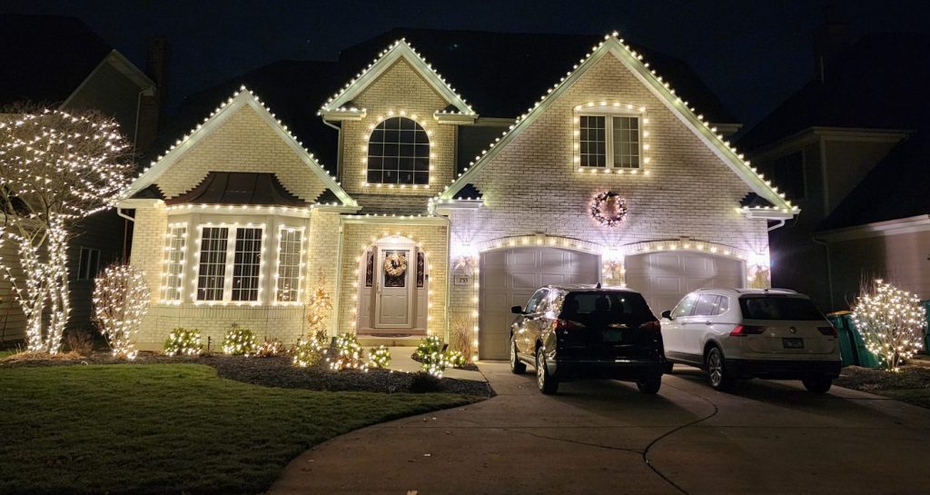 Light brick house with warm white roofline lights on every gable, a lit wreath above the garage and lit shrubs along the walk