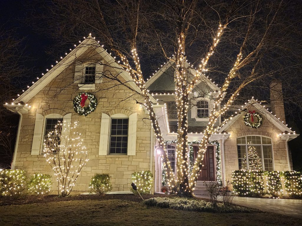 Stone house with warm white roofline lights, two wreaths with red bows, bare trees wrapped branch by branch in warm white lights and lit shrubs along the front