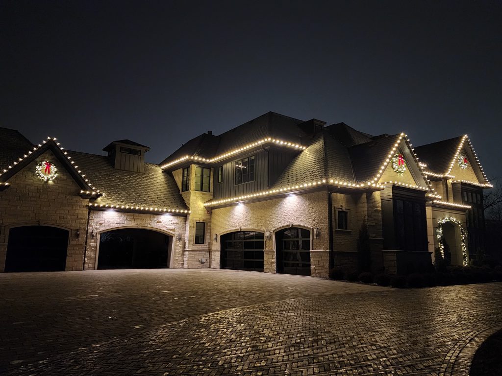 Stone house with four garage bays, rooflines outlined in warm white lights, wreaths with red bows on three gables and a lit garland arch over the entry
