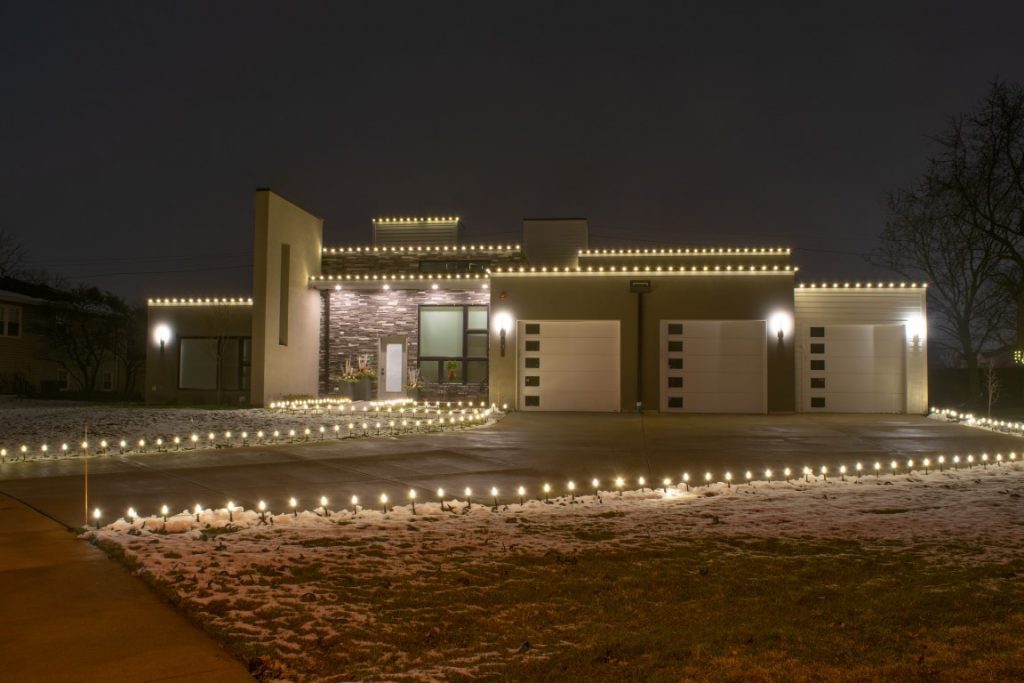 Modern house in light snow with warm white lights along the roofline and a row of small lights on posts along the driveway