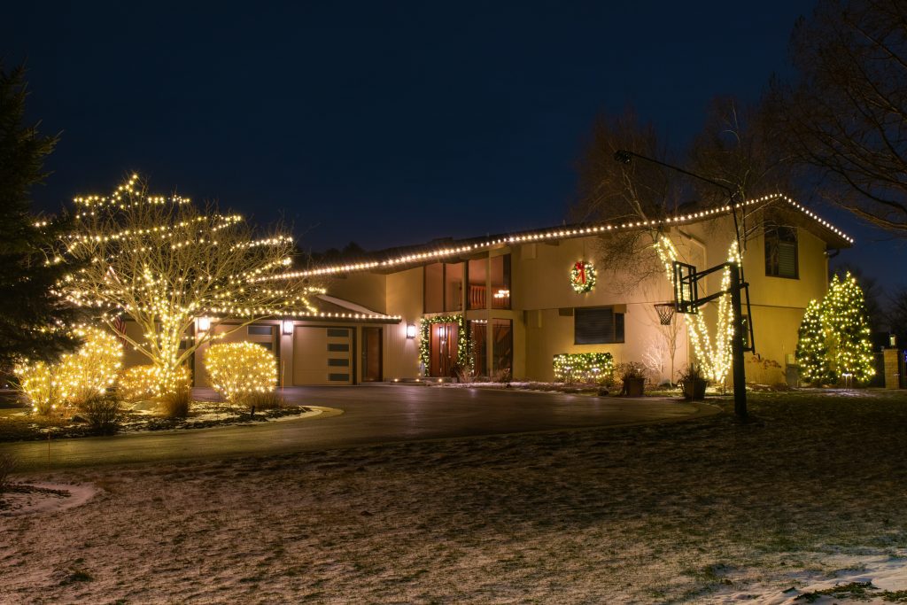 House with warm white roofline lights, trees wrapped in warm white lights and lit shrubs along the drive, light snow on the lawn