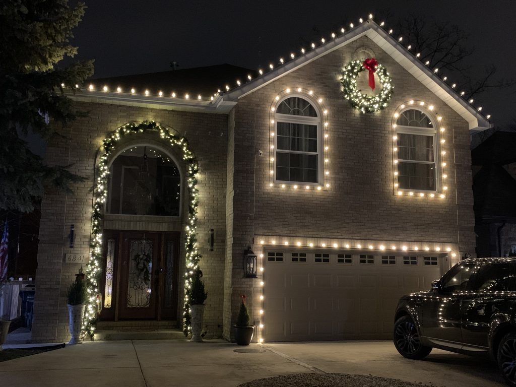 Brick house with warm white lights on the roofline, around two arched windows and over the garage, a wreath with a red bow on the gable and lit garland around the front door