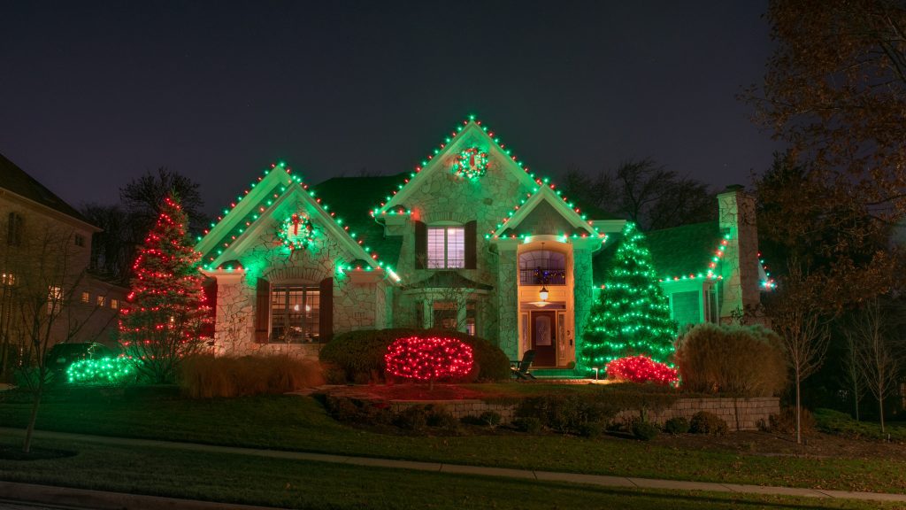 Stone house with green and red roofline lights, wreaths on two gable peaks, and evergreens and shrubs netted in red and green lights