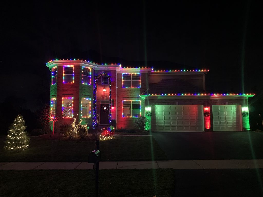 Two-story brick house with multicolor lights outlining the roofline, windows and entry, the garage doors lit in red and green and a small tree wrapped in white lights