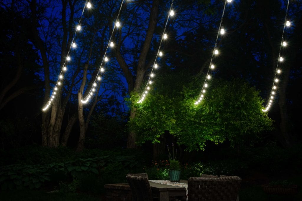 Strings of warm white bistro bulbs hung over a patio with a lit tree and seating below
