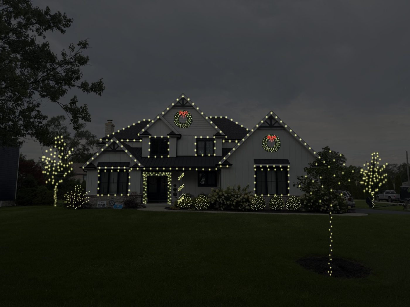 Griswold package example: white house with rooflines, window frames and the entry outlined in warm white lights, two wreaths with red bows, wrapped trees and lit shrubs