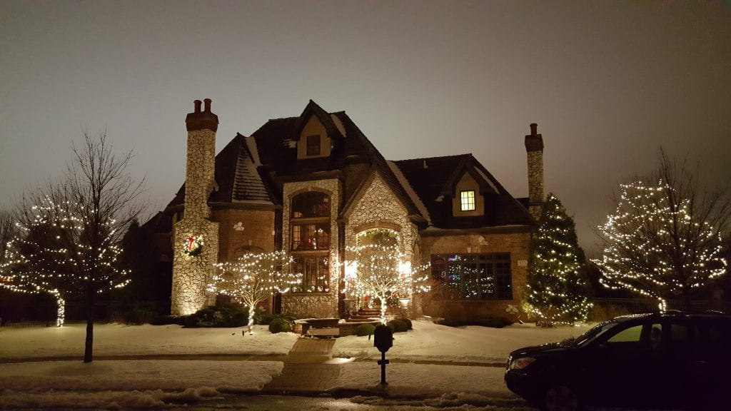 Stone house in the snow with lit trees on both sides of the yard and warm white lights at the front entry