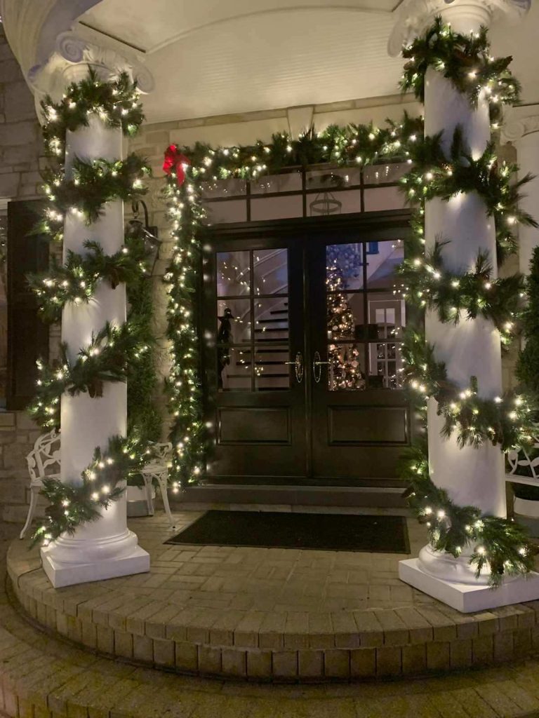 Entry with white columns spiral wrapped in lit garland, more lit garland framing the double doors and a single red bow