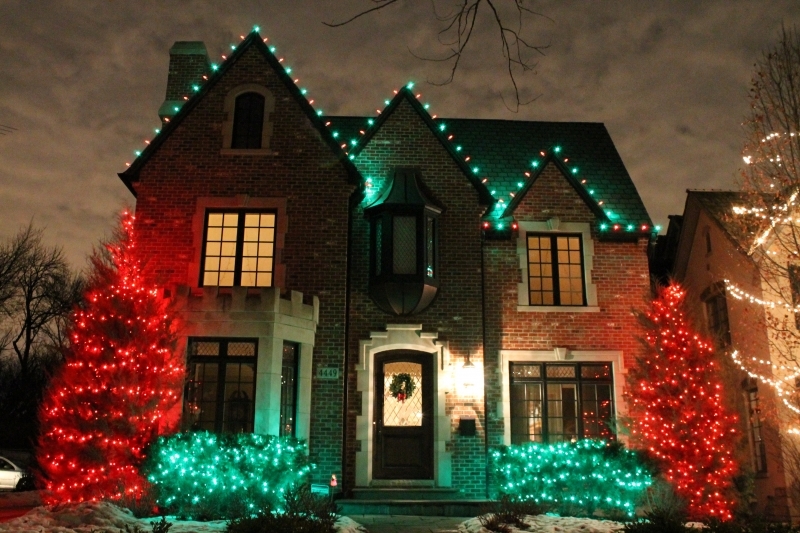 Brick house with green roofline lights, evergreens on both sides covered in red lights and shrubs in green lights