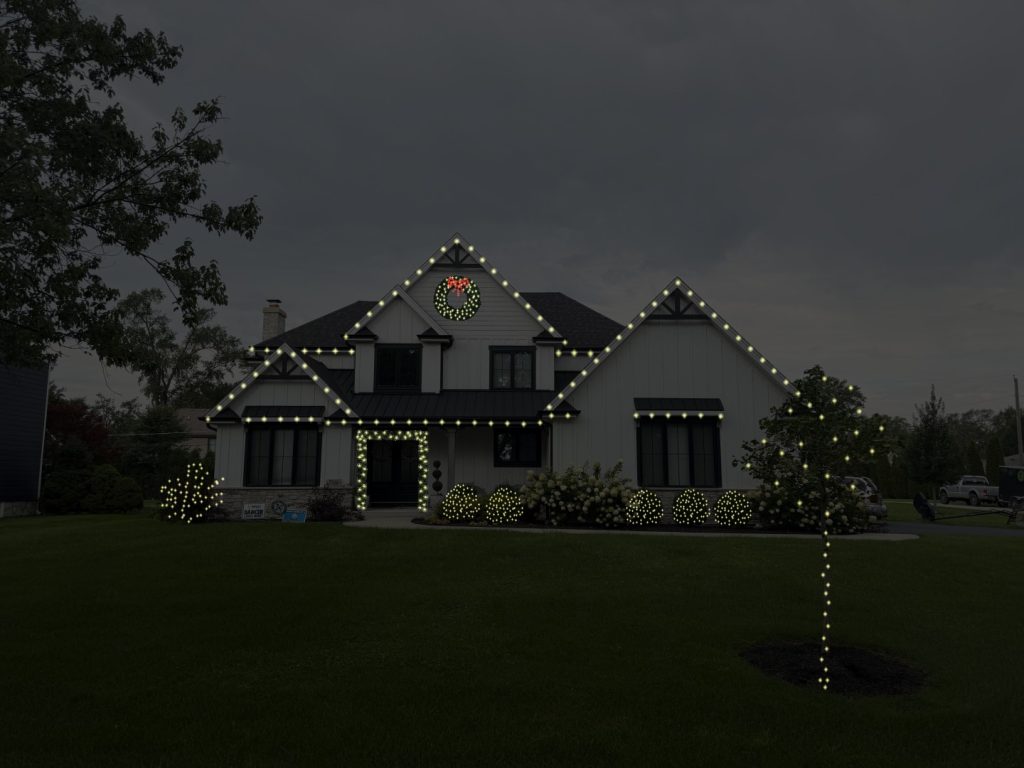 The same house with a lit wreath on the gable, lights framing the front door and a lit tree in the yard