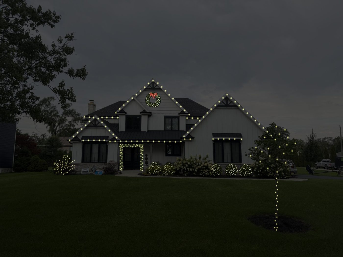 Showstopper package example: white house with the rooflines and entry outlined in warm white lights, one lit wreath, a wrapped tree and lit shrubs