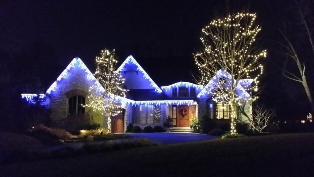 Ranch house with blue and white lights along the roofline and two trees wrapped in warm white lights
