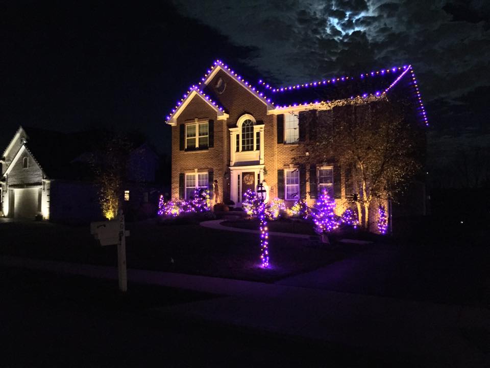 Brick two-story house with purple lights along the roofline and trees and shrubs lit in purple for Halloween