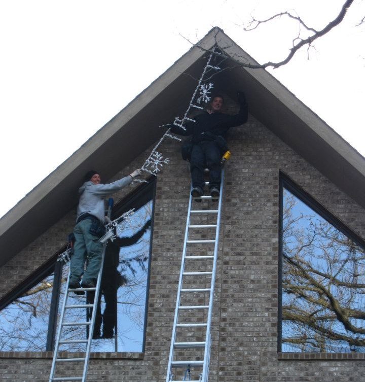 Two installers on ladders hanging lights along the peak of a brick gable